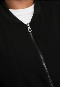 Primo piano di una giacca nera con cerniera sopra una camicia bianca, con particolare attenzione al colletto e al cursore della cerniera con dettagli del marchio.