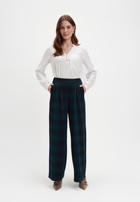 Weiße Bluse mit Knöpfen und Tasche, kombiniert mit hochgeschnittenen karrierten Hosen in dunkelblau und grün. Das Model steht mit den Händen in den Taschen.