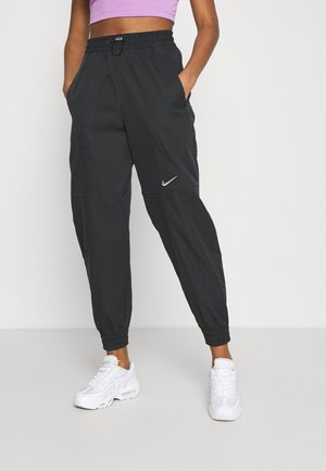 Personne portant un pantalon de jogging noir Nike avec des poignets élastiques et des baskets blanches, debout les mains dans les poches sur un fond uni.