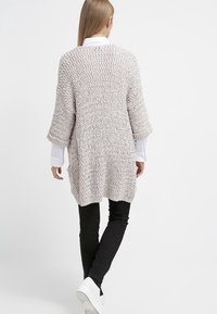 Stickad oversized kofta i ljus beige, med ett kraftigt, strukturerat mönster, tre fjärdedelars ärmar och en avslappnad passform. Matchad med mörka leggings och vita sneakers.