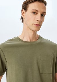 Oliven grøn crew neck t-shirt lavet af blød bomuld. Har kortærmer og en frontlomme på brystet med kontrastsyning.