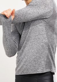 Grijze long-sleeve sportshirt van atletisch materiaal met een textuur, voorzien van duimgaten bij de manchetten en een pasvorm die zich aan het lichaam aanpast.
