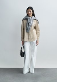 Stripet beige skjorte med knapper, grå genser drapert over skuldrene, hvite bukser med vide ben, holder en liten svart håndveske. Glatte teksturer, moderne design.