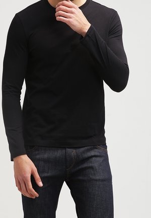 Mand iført en sort langærmet skjorte og mørkeblå jeans, med den ene hånd nær mund og den anden afslappet ved siden.