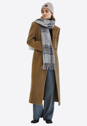 Lange bruine wollen jas, grijs geruite sjaal met franjes, zwarte coltrui, grijze wijde broek, en een lichtgrijze beanie. Platte zwarte schoenen.