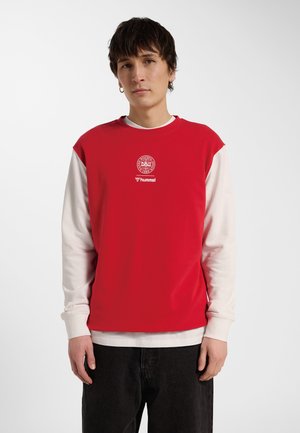 Ung person med kort mørkt hår iført rød ærmeløs overdel over hvid langærmet skjorte og sorte bukser, stående mod hvid baggrund.