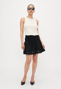 Top cropped de renda floral branca combinado com uma saia preta com elástico, apresentando padrões de ilhós. A modelo usa óculos de sol pretos e sapatos de bico.