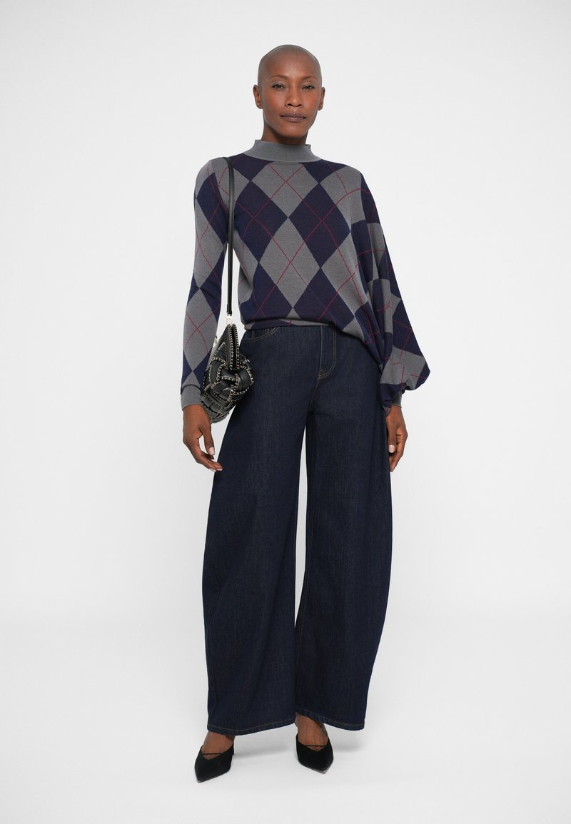 Maglione con motivo argyle grigio, blu navy e rosso; pantaloni a gamba larga in denim scuro; tacchi a punta neri; piccola borsa nera.