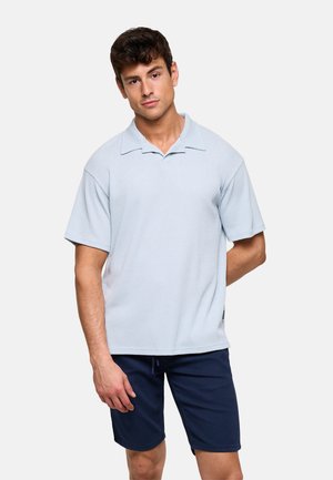 Hombre con camisa de manga corta azul claro con cuello y pantalones cortos azul marino, de pie contra un fondo liso.