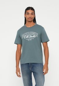 Sinine puuvillane T-särk ümmarguse kaelusega, millel on valge tekst "JACK & JONES Coll. Studio" ja kaunistused. Kandmine siniste teksadega.