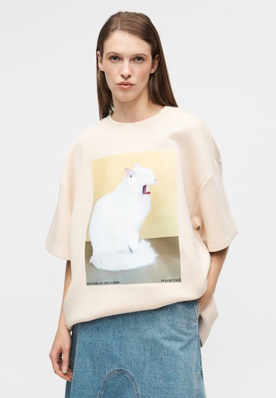 Φαρδιά ροζ ανοιχτό ροζ μπλούζα με λευκό γραφικό γάτας, κοντά μανίκια και εκτυπωμένο σύνθημα στο κάτω μέρος. Φούστα από τζιν ολοκληρώνει την εμφάνιση.