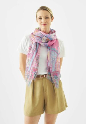 Vrouw draagt een wit T-shirt, beige korte broek met een riem, en een grote roze sjaal met patroon om haar nek gewikkeld, lacht zachtjes.