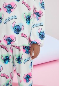 Fehér bolyhos pizsama színes Stitch karakter mintákkal és "OHANA" felirattal, pasztell szív díszítéssel kék, rózsaszín és zöld színekben.