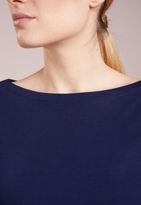 Námořnická modrá vrchní část s lodním výstřihem, žebrovanou strukturou a uvolněným střihem. Tkanina vypadá měkce a pružně, vhodná pro každodenní nošení.