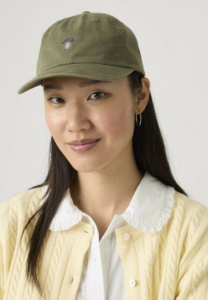Joven con cabello largo y oscuro que lleva una gorra verde oliva con una pequeña flor y un cárdigan crema sobre una camisa blanca con cuello con volantes.