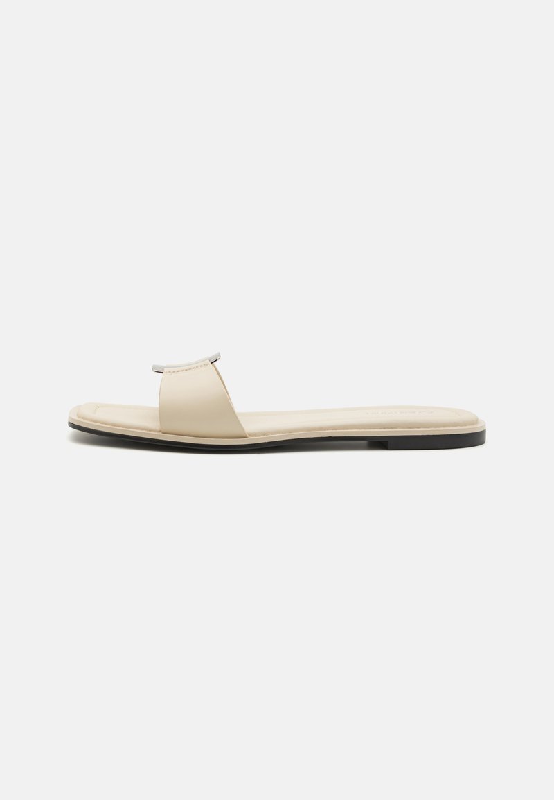 Beige flat sandals with a wide strap and metallic accent. Smooth leather upper, cushioned footbed, and thin black rubber sole.
