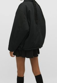 Sort oversized jakke lavet af et glat, mat stof. Har en bred krave, elastisk kant og en subtil glans. Bæres over en plisseret nederdel.