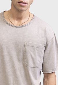 Tricou din bumbac gri deschis, cu gât rotund, mâneci scurte și un buzunar pe piept. Textură moale, cu detalii de cusătură vizibile și croială lejeră.