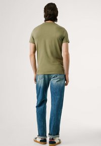 Camiseta verde oliva combinada con jeans de mezclilla azul desgastados y zapatos casuales con acentos en blanco, rojo y azul. Diseño simple, sin patrones.