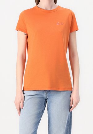 Kobieta ubrana w prostą pomarańczową koszulkę z krótkim rękawem z małym haftowanym napisem na piersi, zestawioną z jasnoniebieskimi dżinsami.