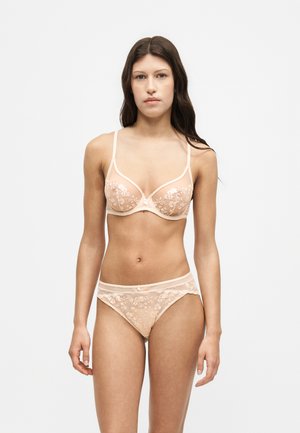 Femme aux longs cheveux bruns, portant un soutien-gorge en dentelle fleurie beige transparent et un sous-vêtement assorti, debout sur un fond blanc uni.