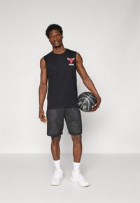 Črna majica brez rokavov z rdečim logotipom Chicago Bulls, v kombinaciji s črnimi mrežastimi kratkimi hlačami in belimi teniskami, drži črno košarko.