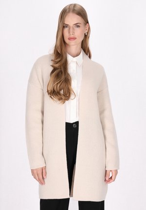 Beige kardigán, nyitott elülső résszel, texturált kötött anyagból készült, hosszú ujja és laza szabása van. Egy fehér galléros ing fölé viselve.