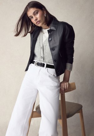 Czarna skórzana kurtka z dużymi guzikami, szary dziergany kardigan, białe szerokie jeansy, czarny pasek oraz drewniane krzesło na neutralnym tle.