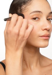Applicazione del trucco con un pennello nero sulla guancia, che mette in mostra un tono di pelle liscio e naturale e un effetto di leggera arrossatura su uno sfondo bianco.