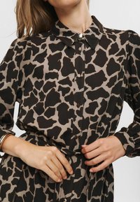 Hnedo-bežové šaty s leopardím vzorom, s golierom, zapínaním na gombíky vpredu a opaskom v páse. Majú dlhé rukávy a riasené manžety. Hladká textúra látky.