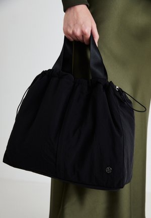 Main de femme tenant un grand sac fourre-tout noir avec de larges anses, sur fond de robe vert olive.