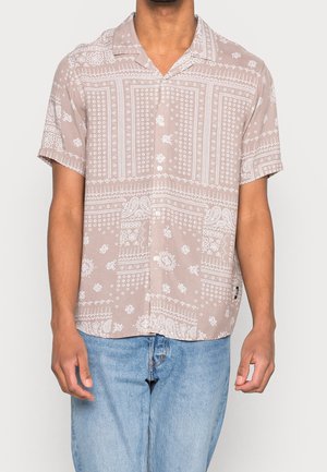Mand iført en beige kortærmet knap-op skjorte med hvide paisley- og blomsterprint, kombineret med lyseblå jeans.