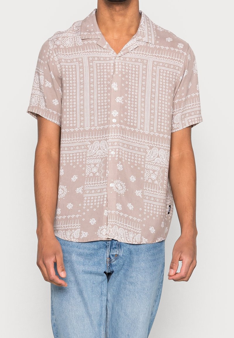 Homme portant une chemise beige à manches courtes boutonnée avec des motifs paisley et floraux blancs, associée à un jean bleu clair.