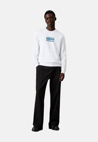 Sudadera blanca con un gráfico azul horizontal en el pecho, combinada con pantalones de pierna ancha negros y zapatos negros. Textura de tela suave.
