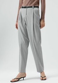 Lichtgrijze geplooide broek met een hoge taille, voorzien van een zwarte riem en een gladde textuur. Getailleerde pijpen, gestyled met zwarte sandalen met hak.