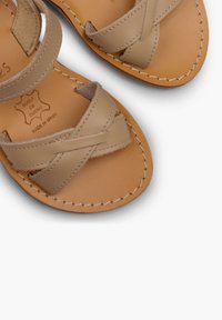 Beige nahkahjulut, joissa on ristikkäiset hihnat, ommeltu yksityiskohtia ja sileä pohja. Sisältää kohokuvioitu tekstin "SUELO DE CUERO" ja "MADE IN SPAIN".