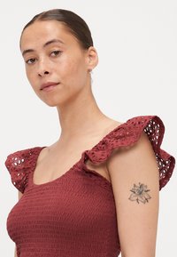 Burgundi värvi smock-kleitus, millel on pitsiline rüütel, tekstureeritud kangas ja avatud mustrid, mida rõhutab broderi lillemotiiv.