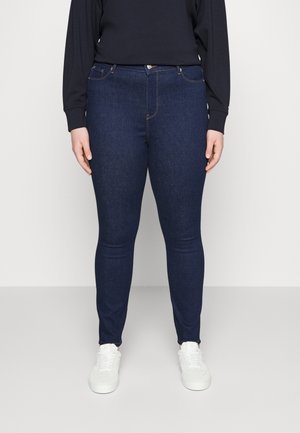 Osoba ubrana w ciemnoniebieskie obcisłe dżinsy, czarną bluzkę z długim rękawem oraz białe trampki, stojąca na jednolitym jasnym tle.