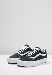 Grüne Wildledersneaker mit weißen Akzenten, flachen weißen Schnürsenkeln, runder Zehenpartie und einer dicken weißen Gummisohle.