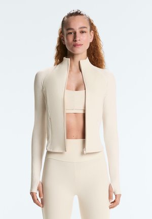 Kvinde med krøllet hår iført en tætsiddende cremefarvet lynlåstrøje, matchende leggings med høj talje og en sports-bh mod en ensfarvet baggrund.