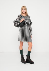 Robe à carreaux noir et blanc avec des manches à volants, cintrée à la taille, associée à des bottes noires à lacets et des chaussettes vertes montantes. Sac à main noir.