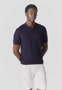 Junger Mann mit kurzen lockigen Haaren, der ein dunkelblaues, kurzärmeliges Strick-Poloshirt und weiße, hochgekrempelte Shorts trägt, vor einem schlichten Hintergrund steht.