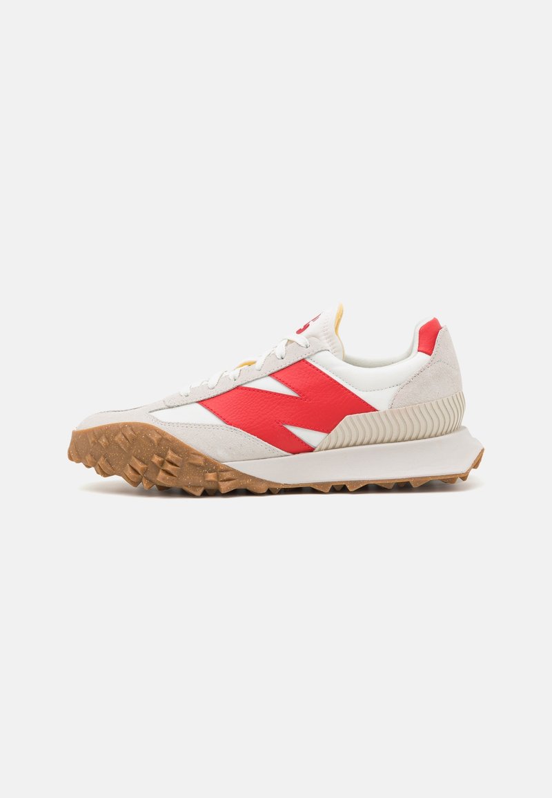 Baskets blanches et beiges avec un logo rouge sur le côté, lacets blancs, semelle en gomme texturée et détail à nervures au niveau du talon sur un fond uni.