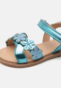 Metallisk teal sandal med blomsterglans, justerbar stropp og buet brun såle. Glatt tekstur med stilig søm.