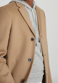 Kameel wollen mengelingjas met gesneden revers, twee zwarte knopen, gedragen over een grijze hoodie. Zachte textuur met een nauwsluitende pasvorm.