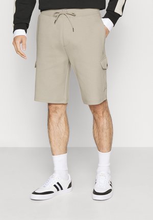 Beige Cargo-Shorts mit Kordelzug an der Taille, getragen von einer Person mit schwarz-weißen Sneakers und weißen Socken vor schlichtem Hintergrund.