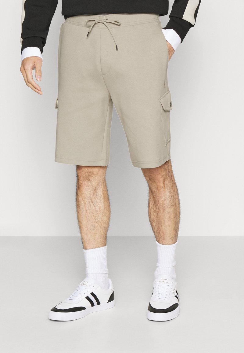 Shorts cargo beiges avec taille à cordon portés par une personne portant des baskets noires et blanches et des chaussettes blanches, sur un fond uni.