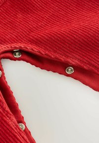 Roter gerippter Stoff mit einer leichten Textur, verziert mit silbernen Druckknöpfen entlang der Kante, zeigt ein geschwungenes Design mit ausgefranstem Saum.