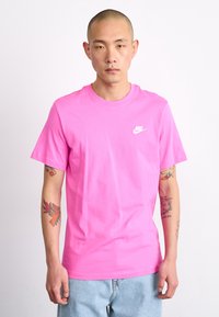 Rožinė medvilninė marškinė su apvaliu kaklu, trumpomis rankovėmis ir mažu baltu Nike logotipu kairėje krūtinės pusėje. Modelis turi tatuiruotes ant rankų.