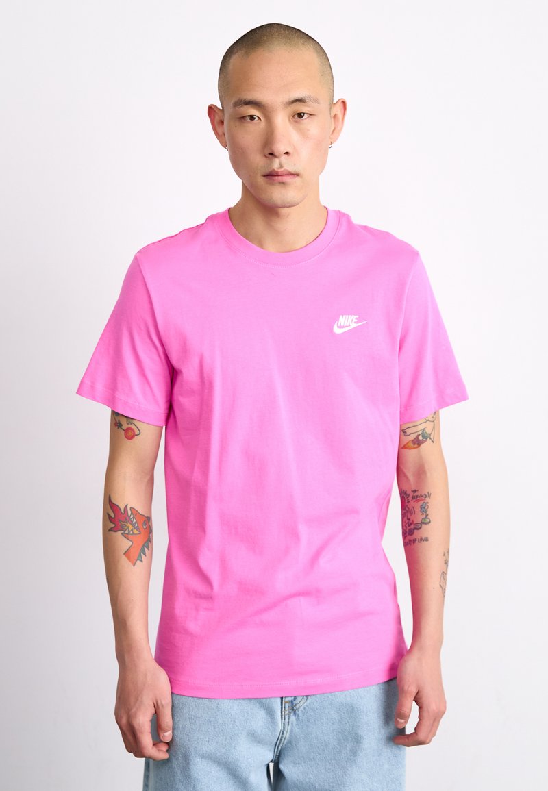 Rožinė medvilninė marškinė su apvaliu kaklu, trumpomis rankovėmis ir mažu baltu Nike logotipu kairėje krūtinės pusėje. Modelis turi tatuiruotes ant rankų.